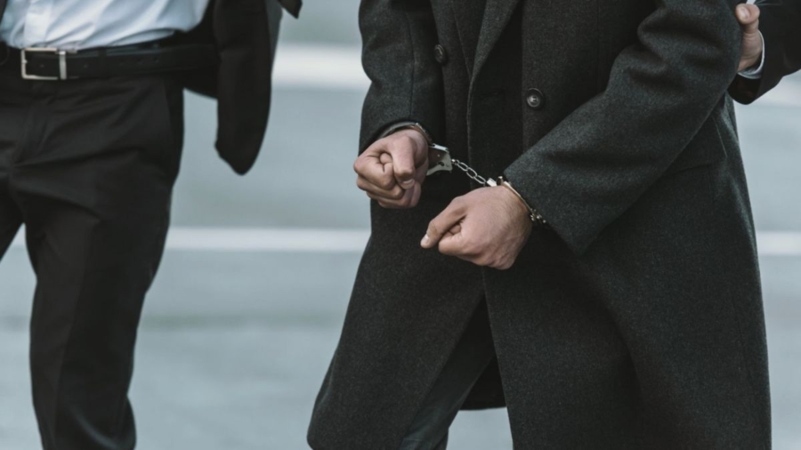 Men dressed in suits walk down the street, with a third man in handcuffs being escorted.