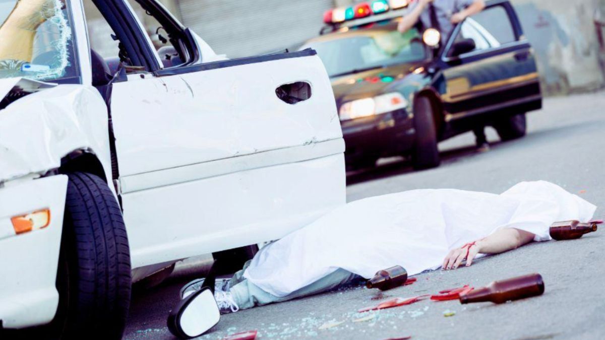 A man lies on the ground beside a car, shattered glass scattered around, indicating a scene of vehicular homicide.