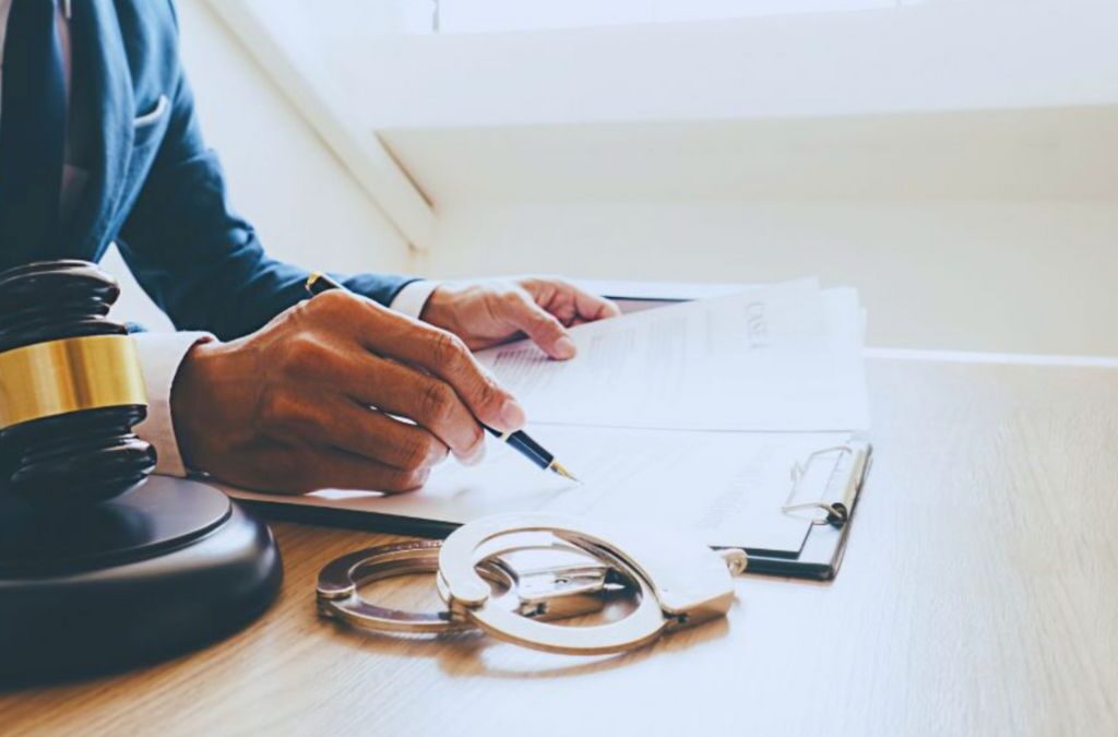 A criminal defense attorney reviews legal documents at a desk, holding a pen. A gavel and handcuffs rest on the desk, representing justice and criminal law.