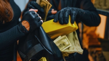 Two masked people wearing black gloves place stacks of cash and a large gold bar into a black bag during a bank robbery, shown in a close-up indoor scene.