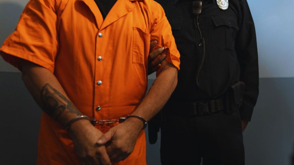 A person in custody stands in restraints beside a uniformed officer in a secure facility.