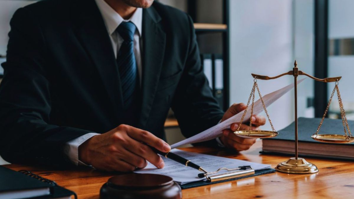 A person in custody stands in restraints beside a uniformed officer in a secure facility.